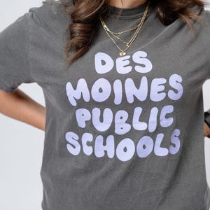 May include: A charcoal grey t-shirt with a purple graphic that reads "Des Moines Public Schools".