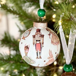May include: A white spherical Christmas ornament featuring illustrations of a nutcracker, a ballerina, a clock, and a rabbit. The ornament has a metallic copper-colored cap, a green wooden bead, and a sheer white ribbon for hanging.