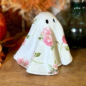 May include: A white ghost figurine featuring a delicate pink rose floral pattern with green leaves. The ghost has two small, dark, oval-shaped eye openings near the top of its rounded head and a draped, flowing shape that reaches the base.