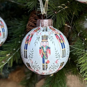 May include: Christmas tree ornaments with a festive nutcracker design. The ornaments are round, white, and decorated with a repeating pattern of nutcracker soldiers in red, blue, and gold. Each ornament has a gold ribbon for hanging.