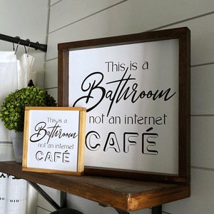 May include: Two wooden signs with black lettering on a white background. The top sign reads "This is a Bathroom not an internet CAFE". The bottom sign reads "This is a Bathroom not an internet CAFE".
