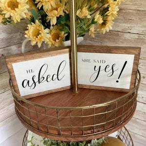 May include: Two wooden signs with the text "He asked" and "She said yes!" on a two-tiered gold wire and wood serving stand. The stand is decorated with greenery and a yellow decorative ball.