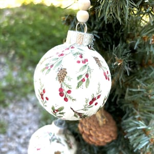 May include: Two white Christmas ornaments with a green and red floral pattern. The ornaments are hanging from a wooden beaded garland on a green pine tree.