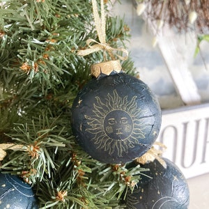 May include: A dark blue Christmas ornament with a gold sun and stars design. The ornament is hanging on a green Christmas tree.