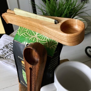 May include: Wooden coffee scoop holder with a scoop, a coffee bag, and a white mug. The holder has a slot for the scoop and a small cup for holding coffee grounds. The coffee bag is labeled "Kona Blend" and "Medium Roast".