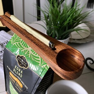 May include: A wooden coffee scoop with a handle that clips onto a bag of Kona Blend coffee. The bag is black with green and white tropical leaf designs and the text "Kona Blend" and "Medium Roast".