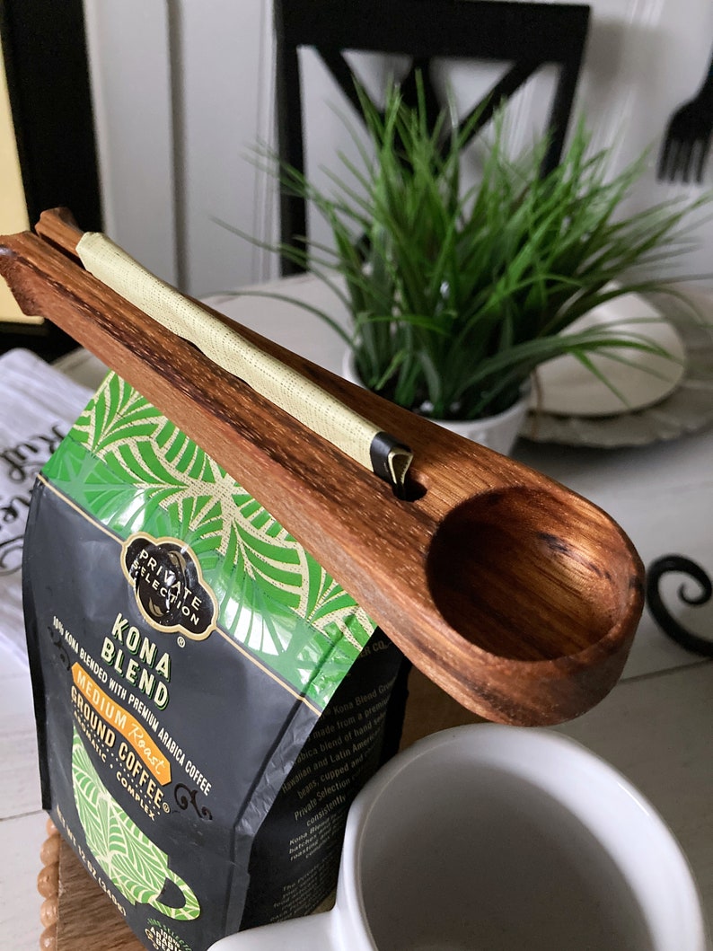 May include: A wooden coffee scoop with a handle that clips onto a bag of Kona Blend coffee. The bag is black with green and white tropical leaf designs and the text "Kona Blend" and "Medium Roast".