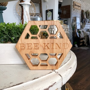 May include: Wooden hexagonal sign with a honeycomb design and the words "Bee Kind" engraved on it. The sign is sitting on a white table.