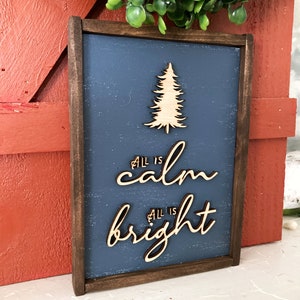 May include: A wooden sign with a dark brown frame and a blue background. The sign features a cutout of a pine tree and the text "All is calm All is bright" in a cursive font.