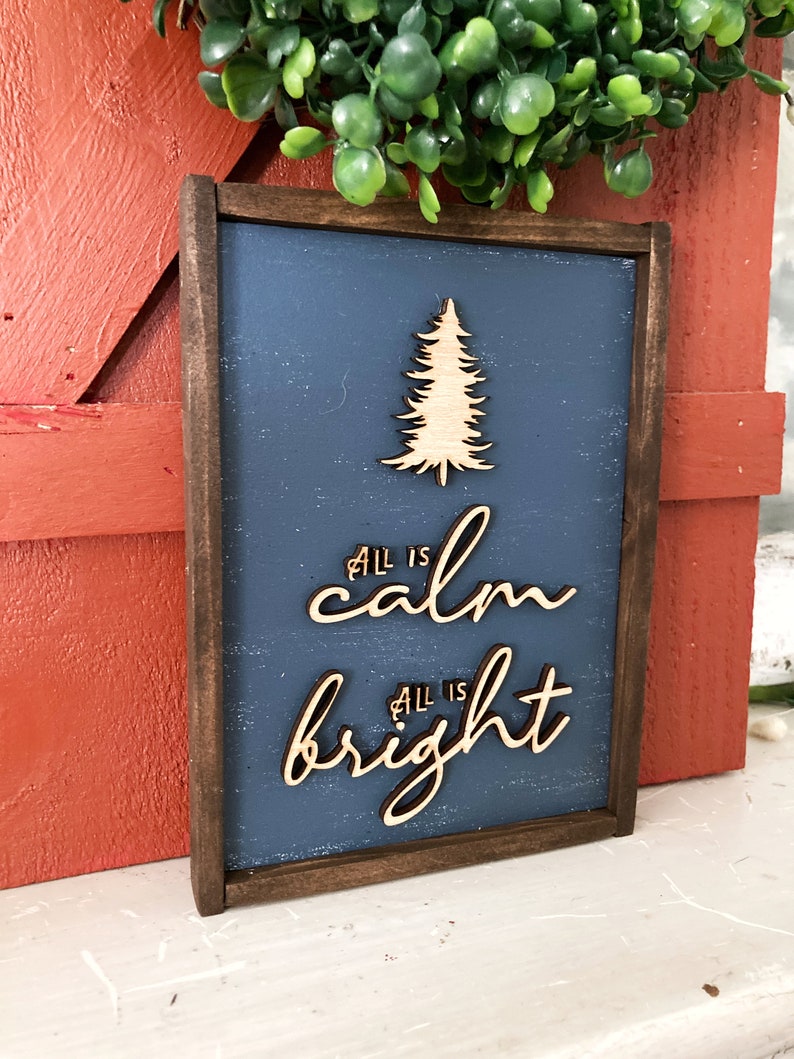 May include: A wooden sign with a dark brown frame and a blue background. The sign features a cutout of a pine tree and the text "All is calm All is bright" in a cursive font.