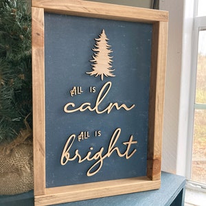 May include: A wooden sign with a dark blue background and a wood-toned frame. The sign features a cutout of a Christmas tree and the text "All is calm, All is bright".