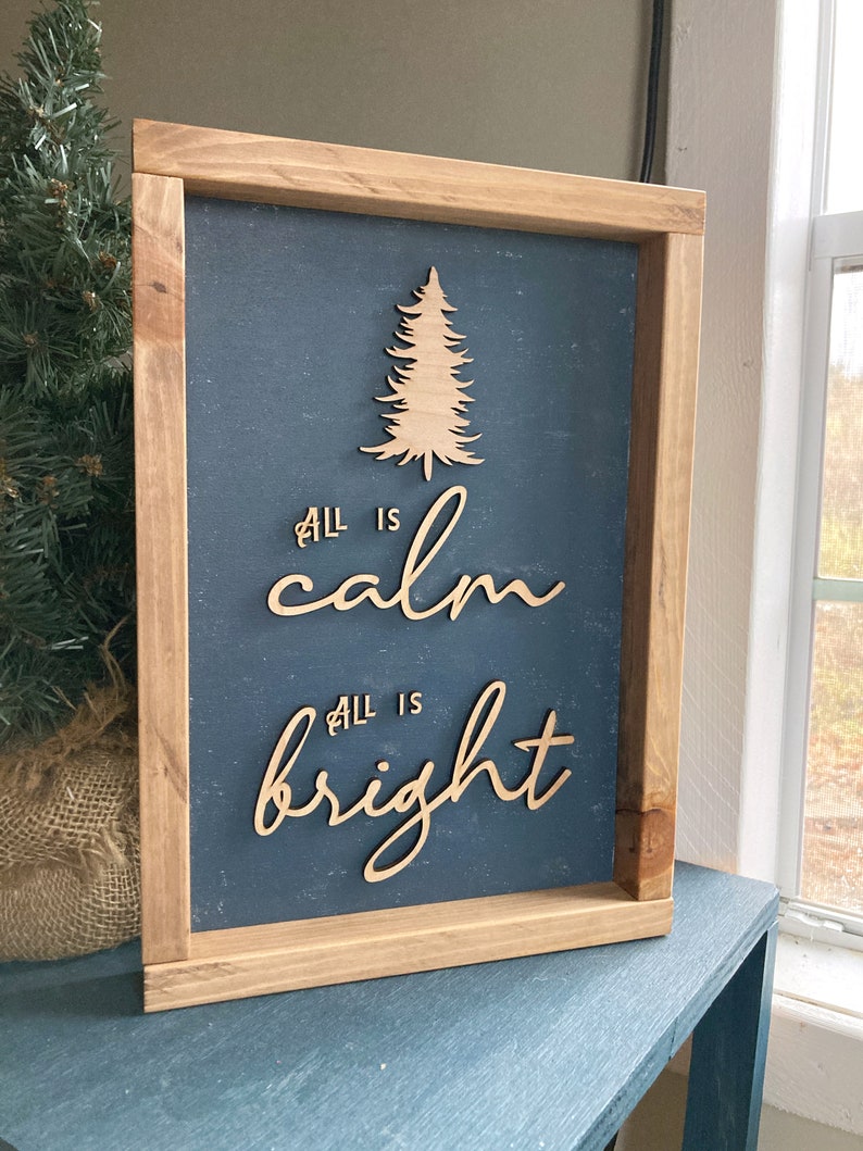 May include: A wooden sign with a dark blue background and a wood-toned frame. The sign features a cutout of a Christmas tree and the text "All is calm, All is bright".