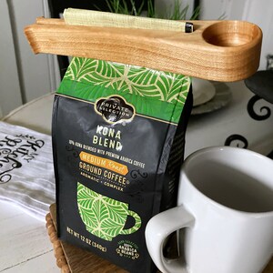 May include: A wooden coffee bag holder with a scoop attached. The holder is holding a bag of Kona Blend coffee. The bag is black with green accents and says "Private Selection Kona Blend 10% Kona Blended with Premium Arabica Coffee Medium Roast Ground Coffee Aromatic Complex Net Wt 12 oz (340g) 100% Arabica Beans".