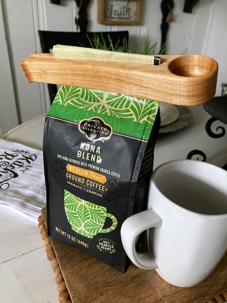 May include: A wooden coffee bag holder with a scoop attached. The holder is holding a bag of Kona Blend coffee. The bag is black with green accents and says "Private Selection Kona Blend 10% Kona Blended with Premium Arabica Coffee Medium Roast Ground Coffee Aromatic Complex Net Wt 12 oz (340g) 100% Arabica Beans".