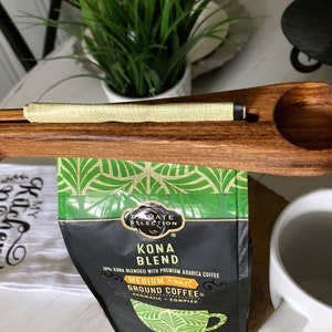 May include: Wooden coffee scoop with a built-in bag clip. The scoop is holding a bag of Kona Blend coffee. The bag has a green and black tropical design with the text "Private Selection Kona Blend 10% Kona Blended with Premium Arabica Coffee Medium Roast Ground Coffee Aromatic Complex".
