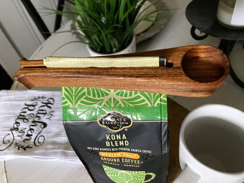 May include: Wooden coffee scoop with a built-in bag clip. The scoop is holding a bag of Kona Blend coffee. The bag has a green and black tropical design with the text "Private Selection Kona Blend 10% Kona Blended with Premium Arabica Coffee Medium Roast Ground Coffee Aromatic Complex".
