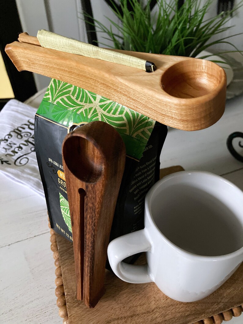 May include: Wooden coffee scoop holder with a scoop, a coffee bag, and a white mug. The holder has a slot for the scoop and a small cup for holding coffee grounds. The coffee bag is labeled "Kona Blend" and "Medium Roast".