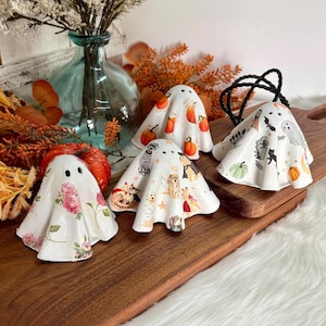 May include: Four white ceramic ghost figurines with varied patterns. One features pink roses, another orange pumpkins, the third various dog breeds, and the fourth a mix of bats, tombstones, black cats, and pumpkins. Each has two small black eye holes.
