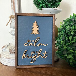 May include: A wooden sign with a blue background and a wood-burned design of a Christmas tree. The sign reads "All is calm, All is bright".