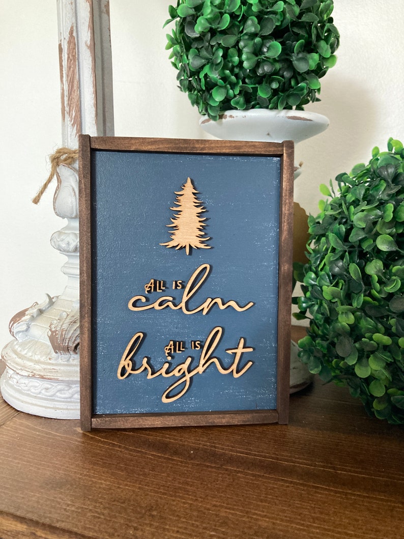 May include: A wooden sign with a blue background and a wood-burned design of a Christmas tree. The sign reads "All is calm, All is bright".