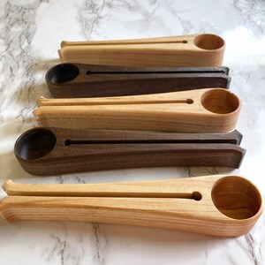 May include: Four wooden coffee scoops in different wood tones. The scoops are long and slender with a rounded end for scooping coffee beans. The scoops are arranged in a row from left to right, with the lightest wood tone on the left and the darkest wood tone on the right.