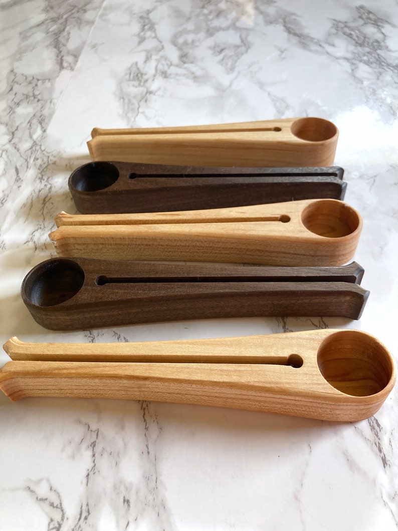 May include: Four wooden coffee scoops in different wood tones. The scoops are long and slender with a rounded end for scooping coffee beans. The scoops are arranged in a row from left to right, with the lightest wood tone on the left and the darkest wood tone on the right.