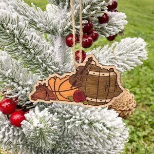 May include: Wooden Christmas ornament shaped like the state of Kentucky with a basketball, a fiddle, a rose, and a barrel. The ornament is hanging on a snowy artificial Christmas tree.