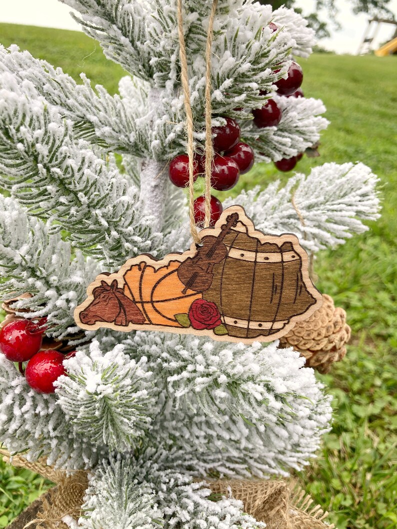 May include: Wooden Christmas ornament shaped like the state of Kentucky with a basketball, a fiddle, a rose, and a barrel. The ornament is hanging on a snowy artificial Christmas tree.