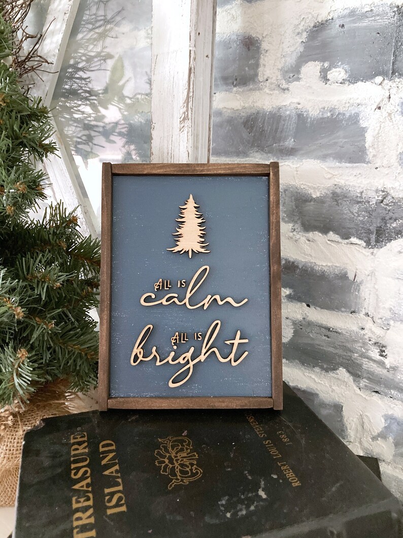 May include: A wooden sign with a blue background and a wooden cutout of a Christmas tree. The sign says "All is calm, All is bright".