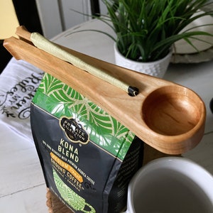 May include: A wooden coffee scoop with a built-in coffee bag holder. The scoop is holding a bag of Kona Blend coffee, which is 100% Kona blended with premium Arabica coffee. The coffee is medium roast and ground.