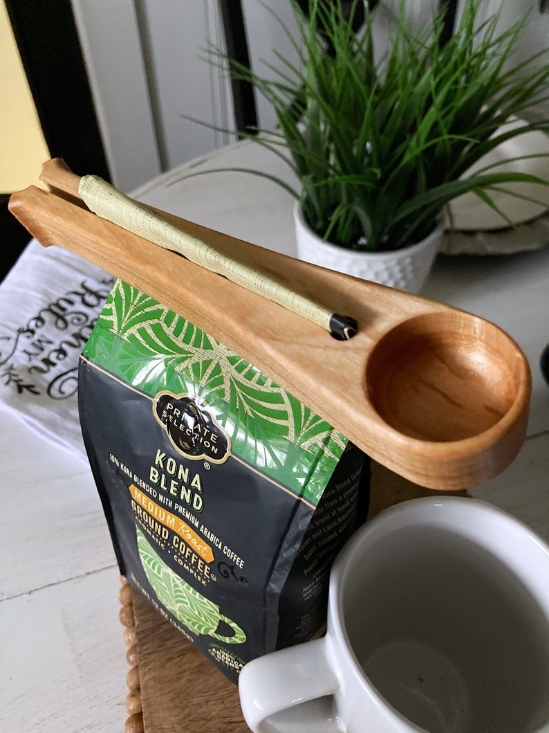 May include: A wooden coffee scoop with a built-in coffee bag holder. The scoop is holding a bag of Kona Blend coffee, which is 100% Kona blended with premium Arabica coffee. The coffee is medium roast and ground.