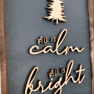 May include: A wooden sign with a gray background and a wood-toned frame. The sign features the text "All is calm, All is bright" in a cursive font. A wood-toned silhouette of a pine tree is above the text.