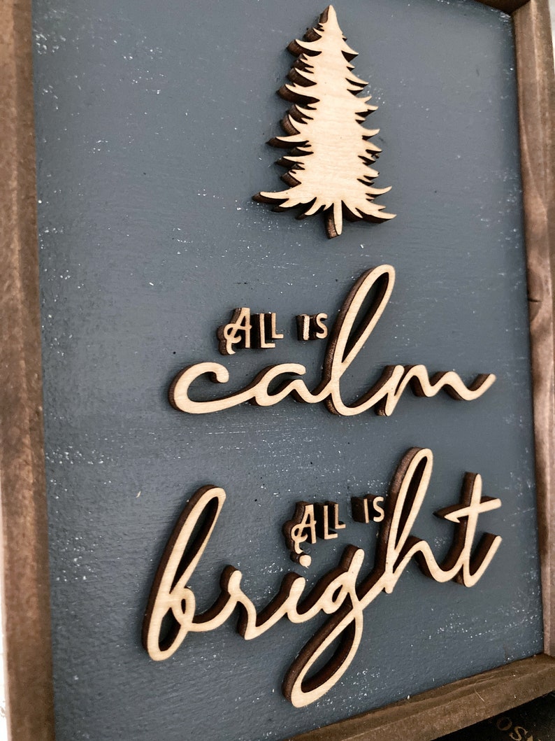 May include: A wooden sign with a gray background and a wood-toned frame. The sign features the text "All is calm, All is bright" in a cursive font. A wood-toned silhouette of a pine tree is above the text.