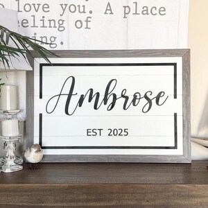 May include: A white wooden sign with a black frame and the text "Ambrose EST 2025" in black lettering.