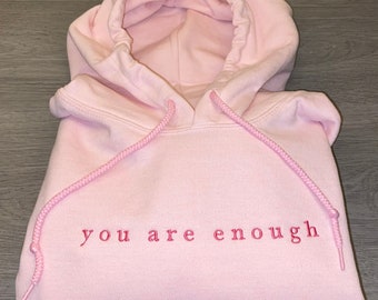 Op de afbeelding: Een lichtroze hoodie met een capuchon met trekkoord. De zin "you are enough" is in bijpassend roze garen op de borst geborduurd. De hoodie is netjes opgevouwen.