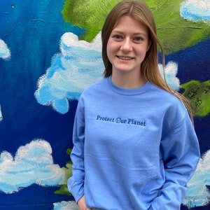 May include: A light blue long-sleeved sweatshirt with the text "Protect Our Planet" and a small globe icon embroidered on the front.