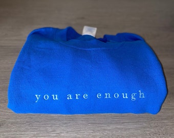 Op de afbeelding: Een gevouwen koningsblauw sweatshirt met de geborduurde tekst "you are enough" in lichtblauwe draad. Het sweatshirt is gemaakt van een zacht materiaal en wordt gepresenteerd op een houten oppervlak.