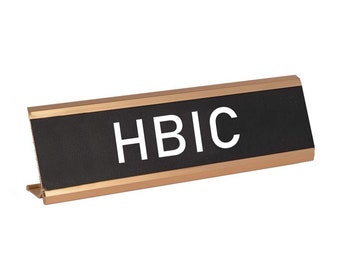 May include: A black desk nameplate with a gold trim and the text "HBIC" in white letters.