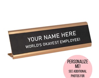 May include: Gold desk nameplate with black background and white text that reads "YOUR NAME HERE WORLD'S OKAYEST EMPLOYEE!"
