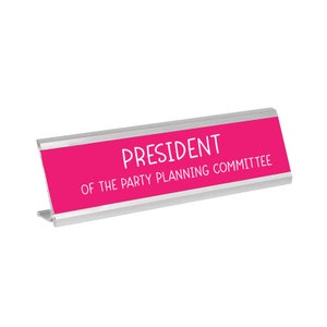 Puede incluir: Una placa de nombre de escritorio de plata con un fondo rosa brillante. El texto en la placa de nombre dice "Presidente del Comité de Planificación de Fiestas."