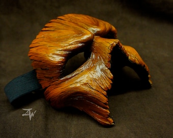 May include: A wooden mask with a textured surface and a dark brown strap. The mask is painted in shades of brown and orange.