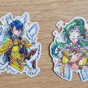 May include: Two stickers featuring anime-style illustrations. One depicts a character in yellow armor with blue hair and white doves. The other shows a character with green hair, holding flowers and a staff. Both are detailed and colorful.