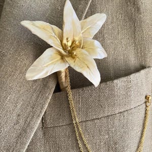 May include: A cream-colored flower lapel pin with gold accents and a gold chain. The flower is attached to a tan jacket pocket. The pin is a stylish accessory for formal wear.