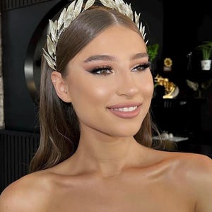 May include: A silver-colored leaf crown, worn on the head. The crown is made of multiple leaves and is worn by a person with long brown hair and makeup. The person is wearing a white strapless top.