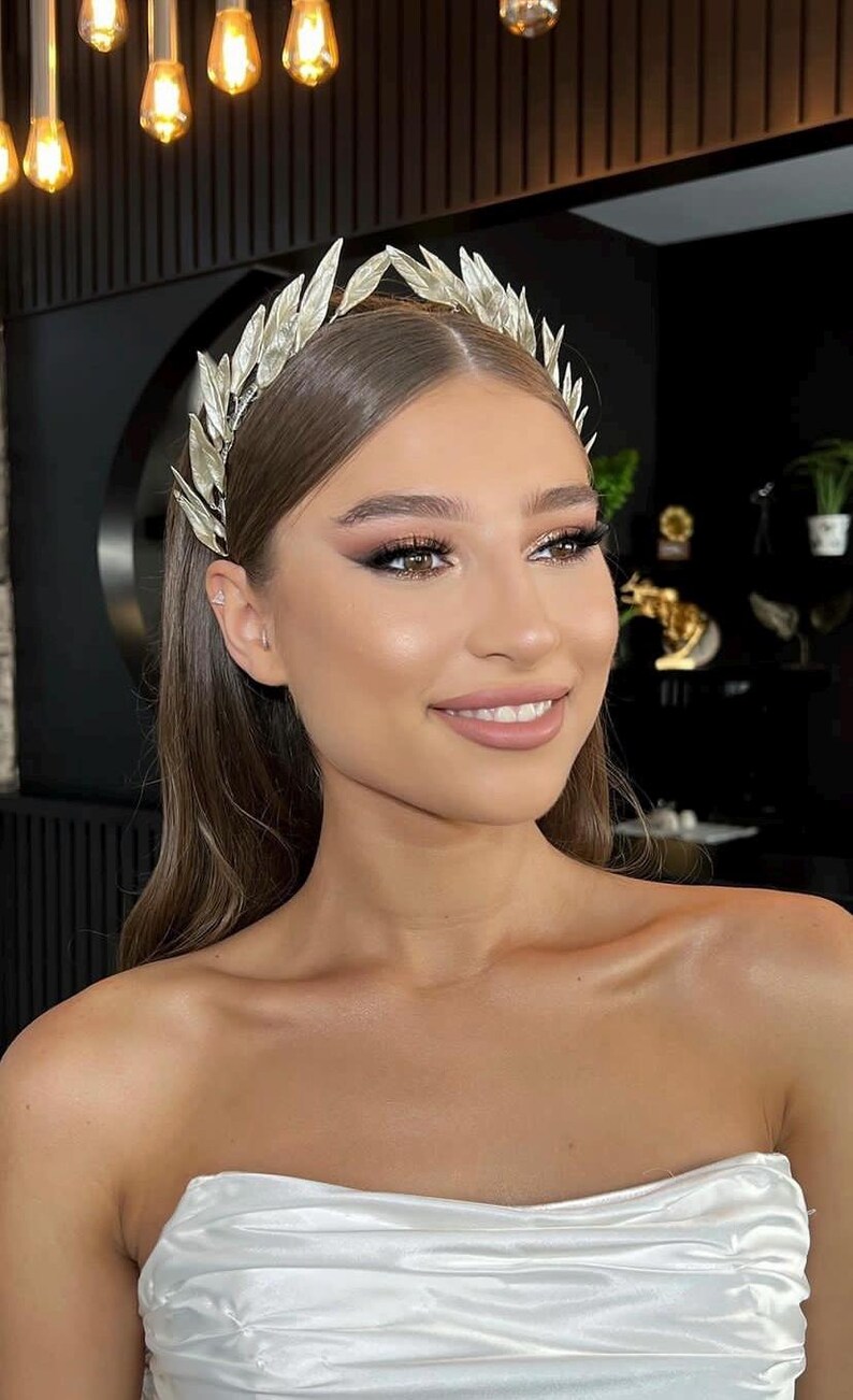 May include: A silver-colored leaf crown, worn on the head. The crown is made of multiple leaves and is worn by a person with long brown hair and makeup. The person is wearing a white strapless top.