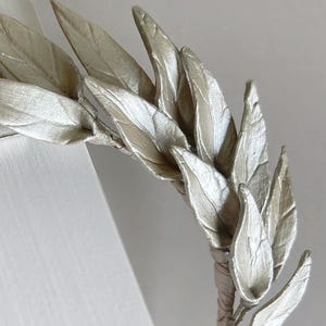 May include: A silver-colored leaf crown, featuring multiple detailed leaves. The leaves are arranged in a circular pattern, with a wrapped stem. The crown is likely for decorative purposes or special events.