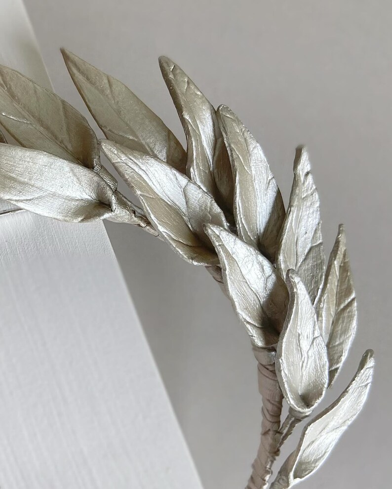 May include: A silver-colored leaf crown, featuring multiple detailed leaves. The leaves are arranged in a circular pattern, with a wrapped stem. The crown is likely for decorative purposes or special events.