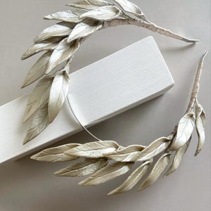 May include: Two decorative leaf hair accessories. Each piece features multiple silvery-white leaves attached to a wrapped stem. The leaves have a subtle sheen, and the stems are wrapped in a neutral-toned material. The accessories are arranged on a white surface.
