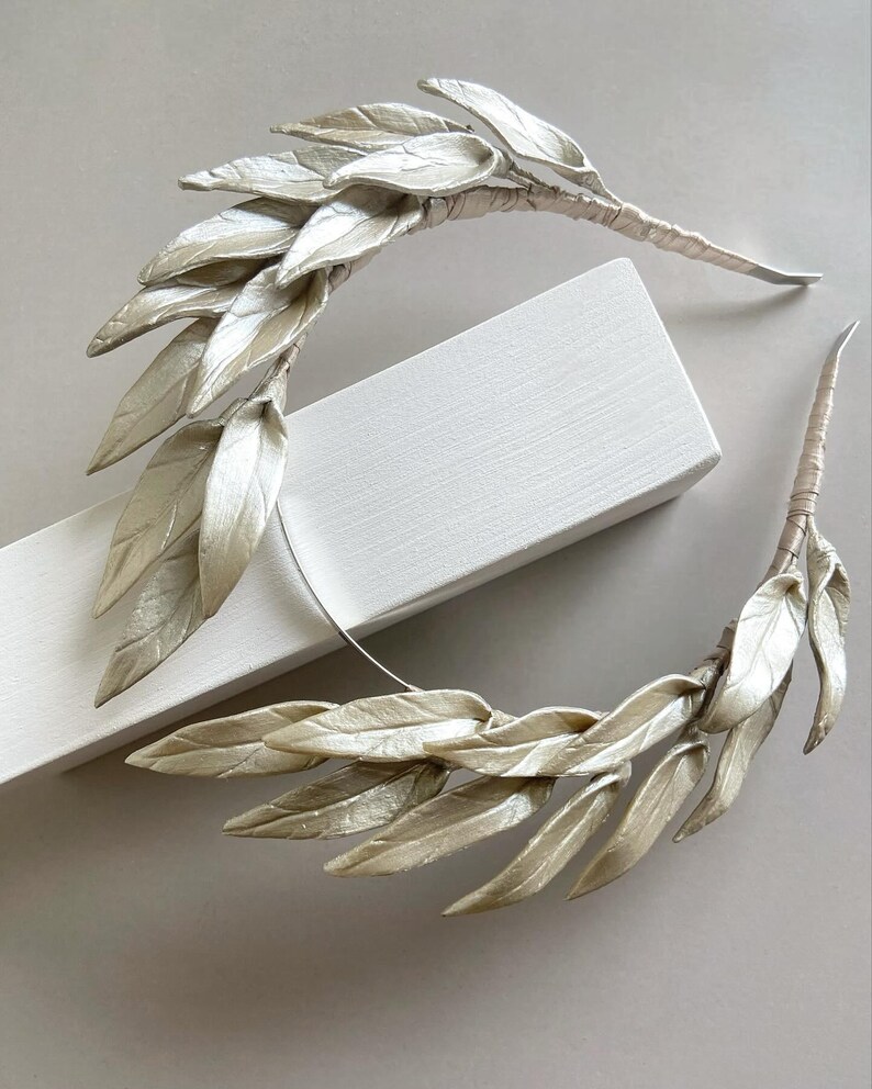May include: Two decorative leaf hair accessories. Each piece features multiple silvery-white leaves attached to a wrapped stem. The leaves have a subtle sheen, and the stems are wrapped in a neutral-toned material. The accessories are arranged on a white surface.