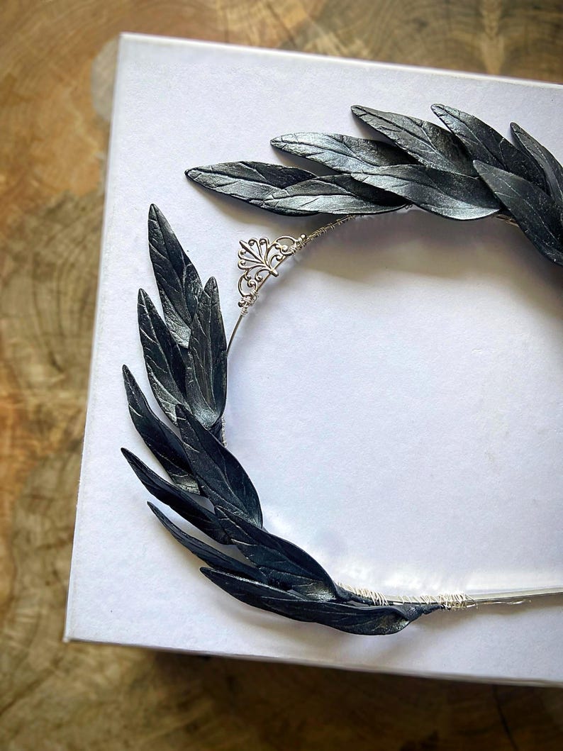 May include: A handcrafted black leaf crown with a silver filigree accent. The crown features dark, textured leaves arranged in a circular design, perfect for a special occasion or costume.